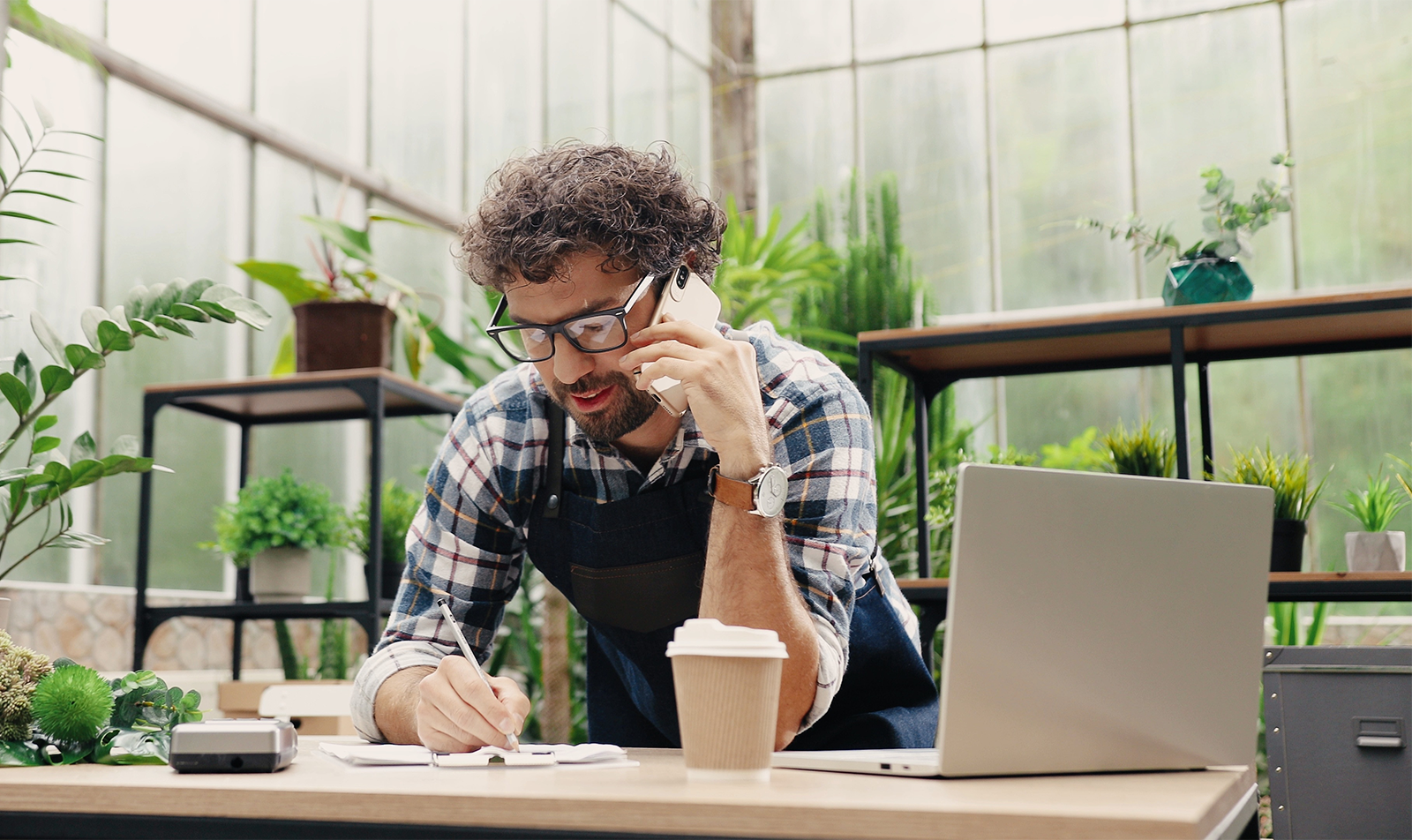 Business owner on the phone taking notes surrounded by plants and a laptop