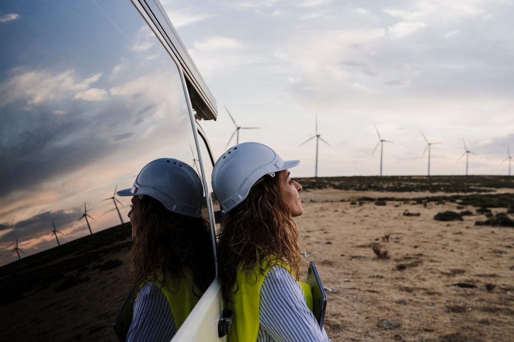 A women wearing a hard hat and high vis leaning against a vehicle with wind turbines in the background