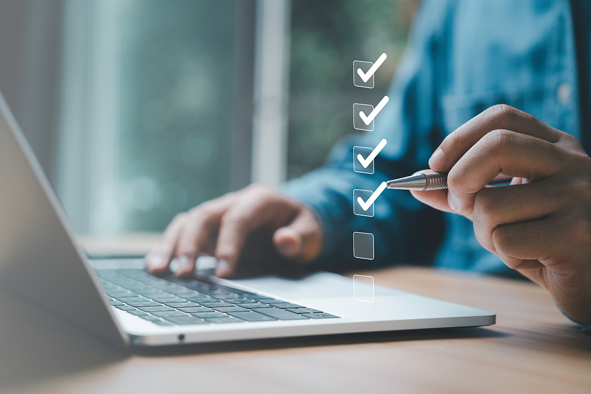 Man sat at laptop at desk and using pen to tick off checklist